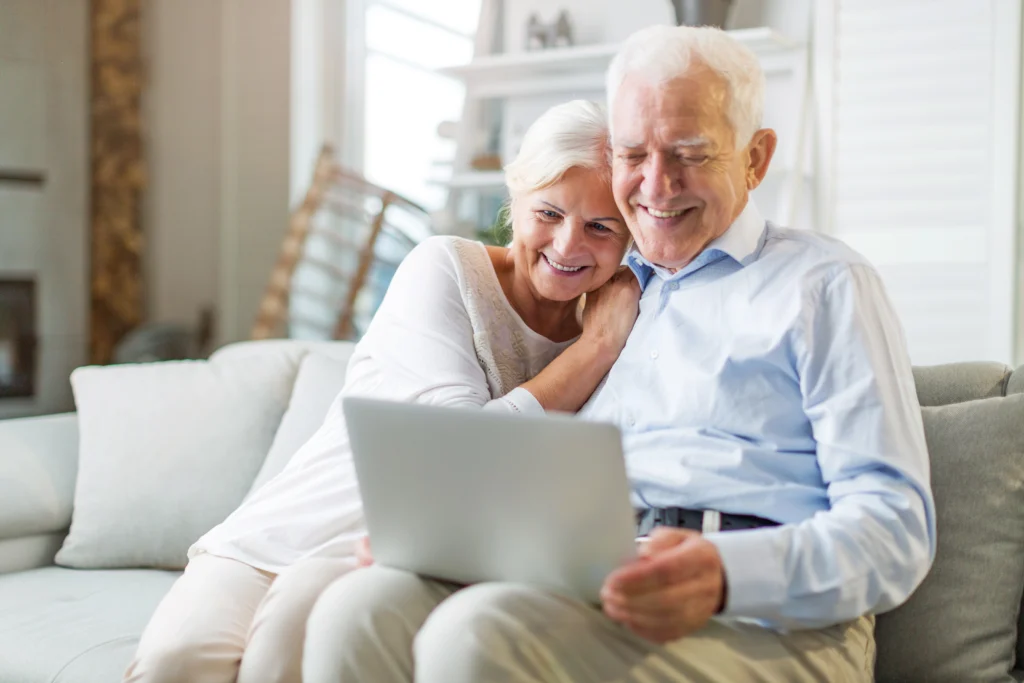 A smiling older couple sit on a couch and look at a laptop together.
