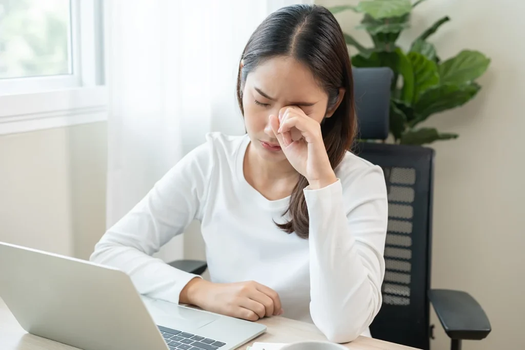 A woman working on a laptop rubs her dry, tired eyes.
