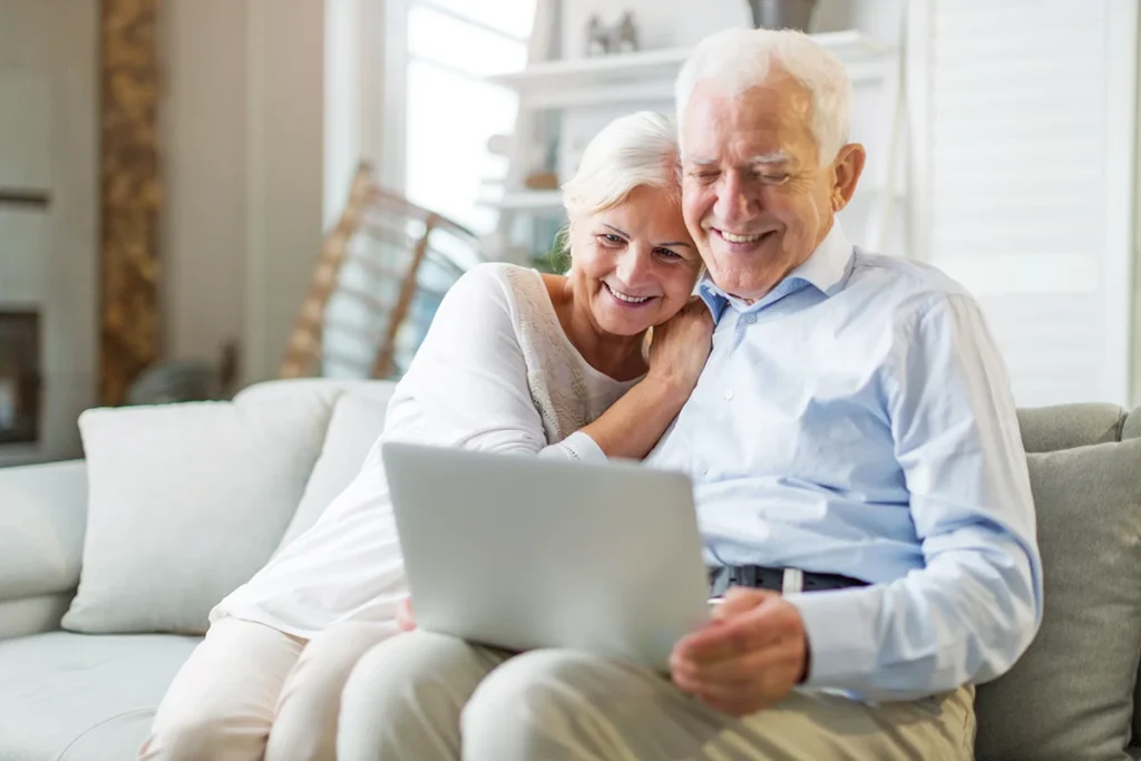 senior-eye-exams A smiling older couple sit on a couch and look at a laptop together.