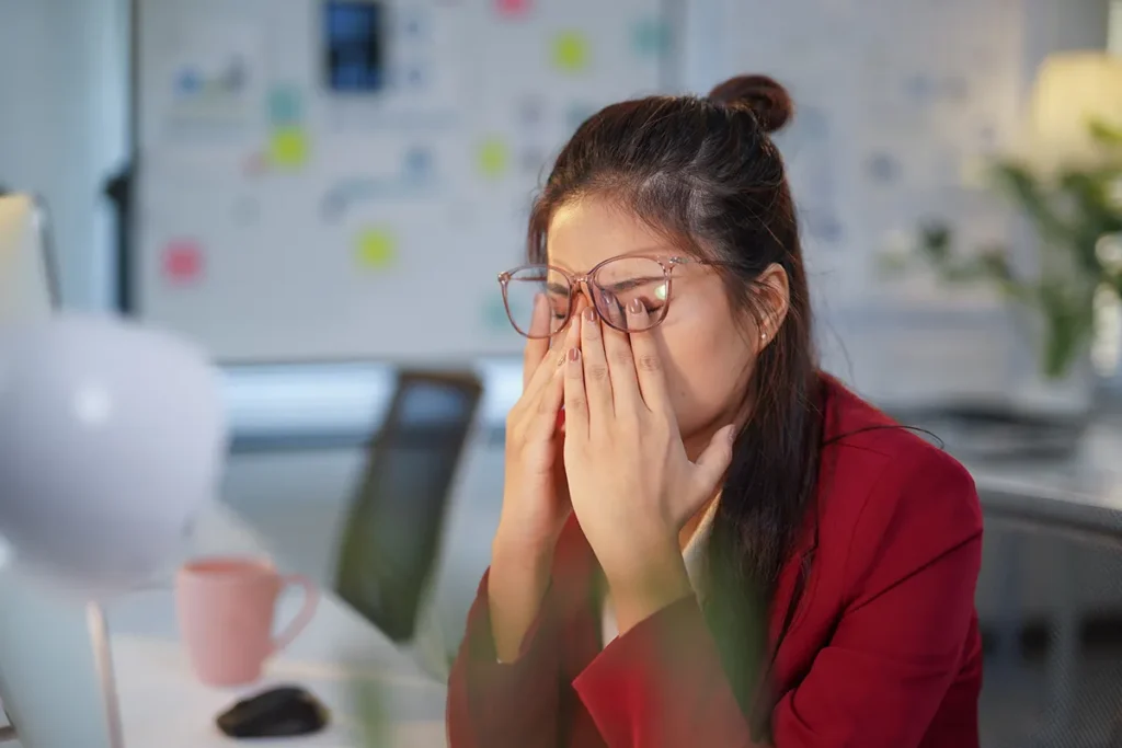 a young woman rubbing her eyes underneath her glasses at work